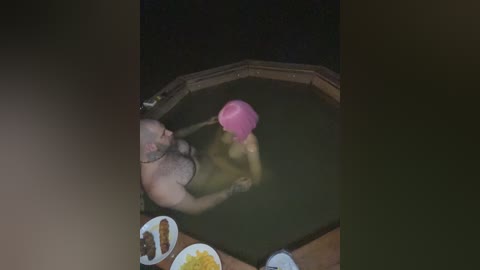 A video of a naked woman in a pink headscarf standing in a bathtub. A man with a bald head and a mustache is behind her, holding her by the waist.