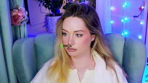 Video of a fair-skinned blonde woman with a light tan, wearing a white dress, sitting in a teal armchair with blue fairy lights and a plant in the background.