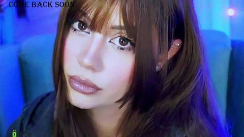A close-up video of a young woman with fair skin, brown eyes, and long brown hair, wearing a dark top, set against a blue and purple background.