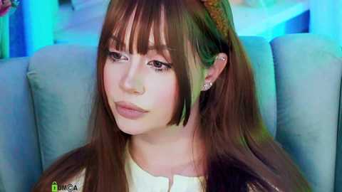 Video of a fair-skinned woman with long brown hair, wearing a white dress, and a gold headband, sitting on a blue sofa, with a colorful, blurry background.