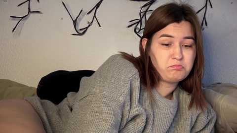 Video of a young woman with straight, shoulder-length brown hair, wearing a gray sweater, lying on a beige couch with a black blanket, against a white wall with abstract black graffiti.