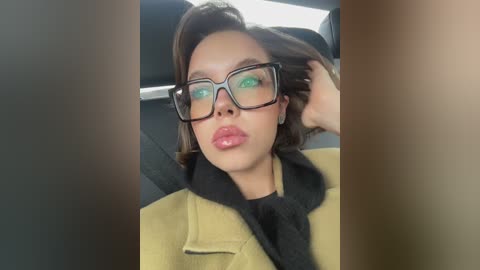 A video of a woman with light brown skin, wearing black-rimmed glasses, a yellow coat, and a black scarf, seated in a car. Her hair is styled in a sleek bob.
