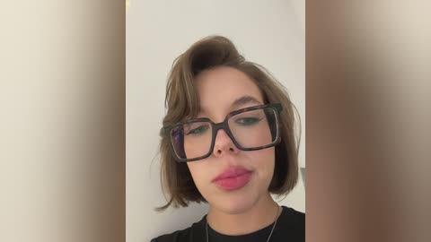 Video of a young woman with light skin, short brown hair, and rectangular black glasses, wearing a black top, against a blurred beige background.