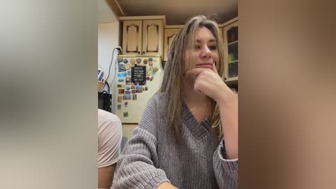 Video of a young woman with long, light brown hair, wearing a grey knitted sweater, sitting in a kitchen. The background features wooden cabinets, a fridge with numerous magnets, and a black cat.