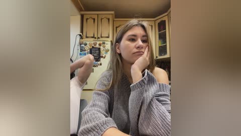 Video of a young woman with light skin, long blonde hair, wearing a grey knit sweater, sitting in a kitchen with wooden cabinets, white refrigerator, and a colorful magnetic board. She is holding a plastic bag and looks contemplative.