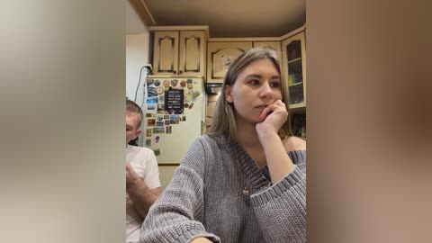 A video of a young woman with light skin, shoulder-length blonde hair, wearing a gray knit sweater, sitting in a cozy kitchen. She gazes thoughtfully, while a man with short hair, wearing a white shirt, stands behind her. The kitchen features wooden cabinets, a fridge adorned with magnets, and a cluttered countertop.