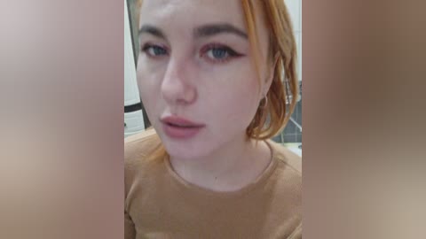 A video of a young woman with fair skin, red hair, and light makeup, wearing a tan shirt. Her expression is neutral.