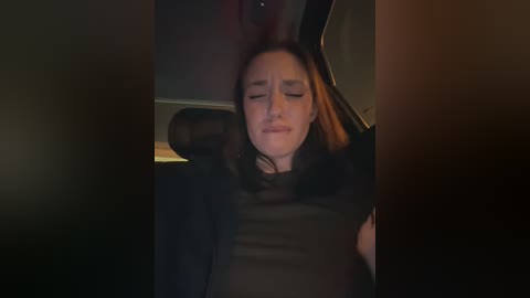 A dimly lit video of a young woman with fair skin and long, dark hair, wearing a black turtleneck, asleep in a car with a blurred, shadowy background.