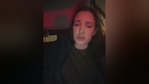 A dimly-lit video captures a woman in a dark, possibly car interior, wearing a black coat, looking exhausted with closed eyes, and a red light illuminates her face.