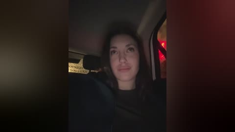 A video of a smiling woman with long dark hair in a dimly lit car. She is wearing a black top, and her face is slightly out of focus.