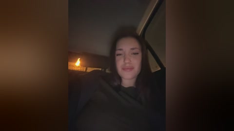 A dimly lit video of a young woman with long, dark hair, wearing a black jacket, smiling gently. A warm, flickering candle glows in the background.