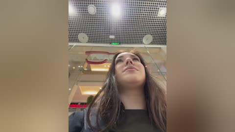 A video of a young woman with long brown hair, wearing a black top, standing in a modern shopping mall. The ceiling features a grid pattern with recessed lights, and the background includes glass panels and soft lighting.