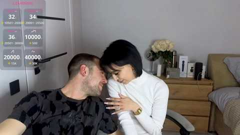 Video of a smiling Asian woman in a white sweater, seated on a male's lap in a hospital bed. He wears a black shirt, and a medical chart displays vital signs.