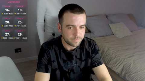 A video of a young man with short brown hair and a beard, wearing a black shirt, sitting on a bed in a modern bedroom with gray bedding and a white wall. A digital display with stats and a leaderboard is overlaid.