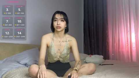 A video of a young Asian woman with short black hair, sitting cross-legged on a bed, wearing a gold sequin halter top and black shorts. The room has a white wall, a calendar, and pink curtains.
