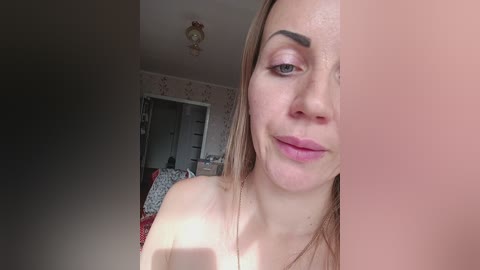 Video of a smiling, fair-skinned woman with blonde hair, wearing pink lipstick, in a bedroom with patterned wallpaper, cluttered with clothes and a bed.