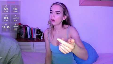 Video of a young Caucasian woman with light skin and long blonde hair, wearing a blue tank top, pointing, indoors with a purple background, bookshelf, and digital fitness tracker overlay.