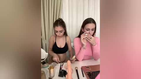 Video of two young women with fair skin, wearing black and pink tops, sitting at a vanity table. One applies makeup while the other holds a phone. Makeup brushes, a dildo, and a mirror are on the table.