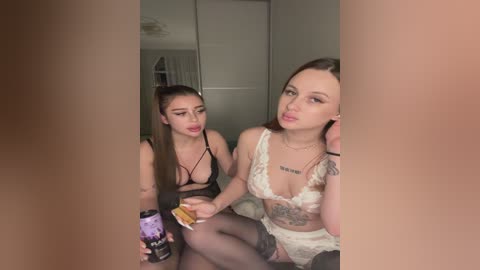 Video of two young women in lingerie, one with a tattoo on her chest, sitting in a dimly lit room, drinking from a can, looking pouty.