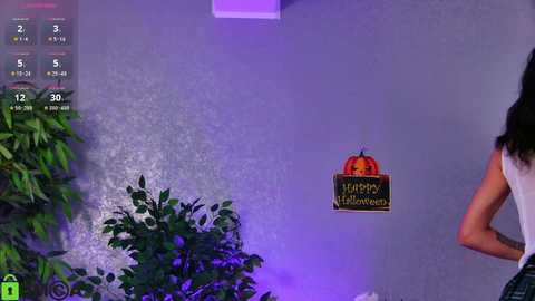 Video of a woman with long black hair in a sleeveless top, standing in front of a gray wall adorned with a \"Happy Halloween\" sign and a potted plant.