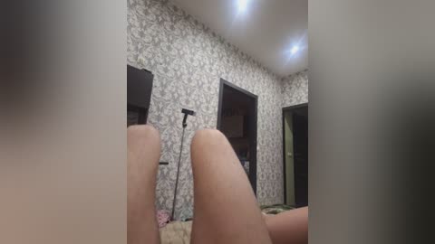 A video shows a dimly lit bedroom with wallpaper featuring a floral pattern. Two legs, presumably male, are visible in the foreground. The room has a TV, a lamp, and a mirror.