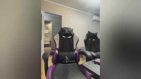 Video of a modern gaming setup with two identical black gaming chairs with purple accents and headrests, positioned in a well-lit room with beige walls, a door, and a small desk visible in the background.