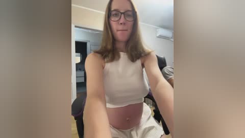 Video of a white woman with straight, shoulder-length brown hair, wearing black-rimmed glasses and a white crop top, sitting in a modern room with a black office chair and white walls.