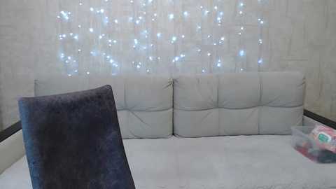 Video of a minimalist living room with a white tufted leather sofa, navy velvet pillow, and a string of blue and white fairy lights on a white brick wall.