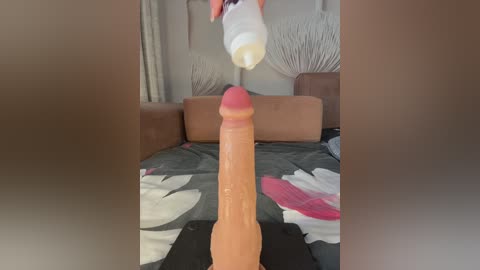 Video of a large, erect, flesh-colored dildo with a realistic texture and detailed veins, positioned on a bed with a floral-patterned black and white comforter. A white baby bottle hangs above the bed, suspended by a string. The background features a beige wall with a macrame hanging.
