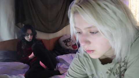 Video of two young women with long hair, one blonde, one brunette, in a dimly lit bedroom. The blonde sits on a bed with a patterned blanket, looking contemplative, while the brunette sits nearby, wearing glasses and a black top.