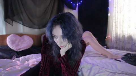 Video of a pale-skinned woman with long, dark hair, lying on a bed in a dimly lit room, wearing a red velvet dress, with a heart-shaped pillow beside her.