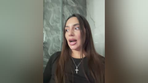 Video of a woman with long brown hair, wearing a black top and necklace, standing in a dimly lit room with a stone wall and blurred background.