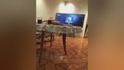 A video of a dimly lit room with a TV displaying a woman, a table with a floral tablecloth, and a wooden floor with geometric patterns.