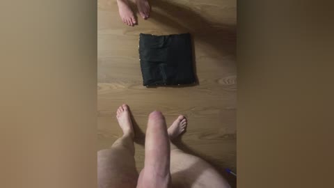 Video of a man's legs and feet on a wooden floor, with a pair of black pants laid out on the floor in front.