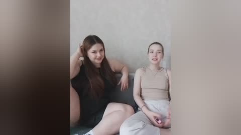Video of two young women, one with long brown hair in a black dress, the other with blonde hair in a beige top, sitting on a grey couch against a beige wall.
