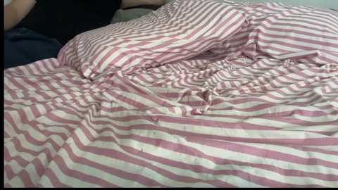 Video of a person sleeping on a bed with pink and white striped sheets, partially covered by a dark shirt. The bedding appears rumpled and disheveled.