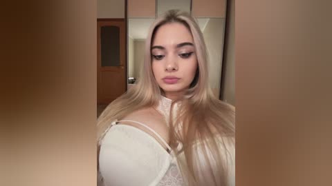 Video of a young Caucasian woman with long, straight platinum blonde hair, wearing a white lace bra with visible cleavage, standing in a beige-walled bedroom with a wooden door and mirrored closet in the background.