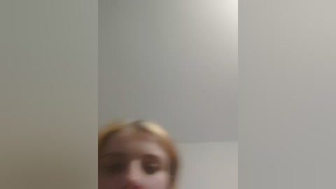 A blurry video of a woman with blonde hair, possibly in a bathroom, with a plain white wall and a subtle light source in the upper right.