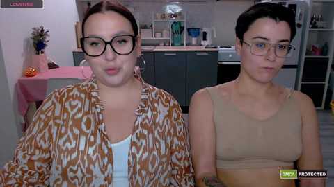 Video of two women in a modern kitchen. One with glasses and patterned shirt, another with short hair and beige top. Background includes cabinets, utensils, and a pink table.