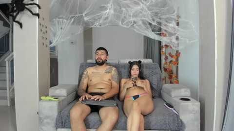 Video of a shirtless tattooed man and a nude woman with black hair and cat ears, sitting on a gray couch in a modern room with white walls and spider web decor.