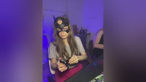 Video of a woman with long brown hair, wearing a black cat mask and a gray shirt, sitting at a dark table, holding a black object, in a dimly lit room with purple lighting.