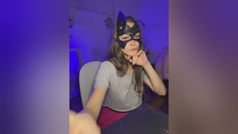 Video of a young woman with long brown hair wearing a Batman mask, gray top, and red skirt, seated indoors under purple lighting, hands on her face.