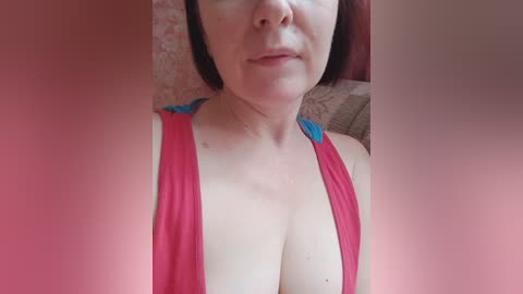 A video of a light-skinned woman with auburn hair, wearing a low-cut red tank top that reveals ample cleavage, seated against a patterned wall.