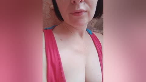 Video of a pale-skinned, short-haired woman in a low-cut pink tank top, showing ample cleavage. The background is a textured beige wall, partially obscured by a pink blur.