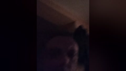 A blurry, dimly lit video shows a person with dark hair, possibly female, facing away from the camera, in a shadowy room with a textured wall and indistinct background.