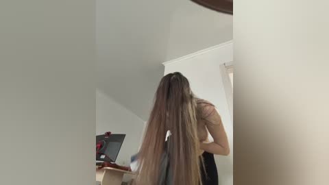 Video of a woman with long, straight brown hair, seen from behind, standing in a sparsely furnished room with a TV and a wooden desk in the background.