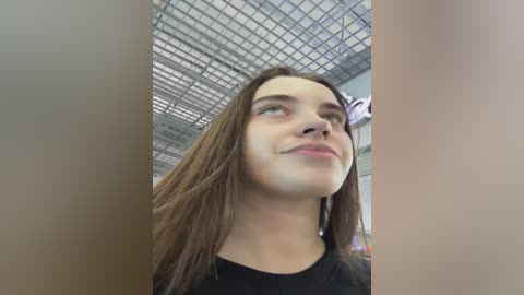 A video of a young woman with long brown hair, light skin, and a subtle smile, taken indoors with a grid ceiling and fluorescent lighting. She wears a black top.