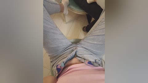 Video of a person's legs wearing light blue jeans with a white shirt, seated on a teal chair, with beige floor tiles visible in the background.