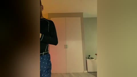 Video of a man's back in a dimly lit room, wearing a black long-sleeve shirt and blue pajama pants with white stars. A beige wardrobe and a white chair are visible in the background.