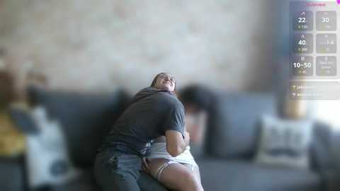Video: A man in a grey shirt and jeans embraces a woman in a white dress on a grey couch, blurred background with a digital timer screen.